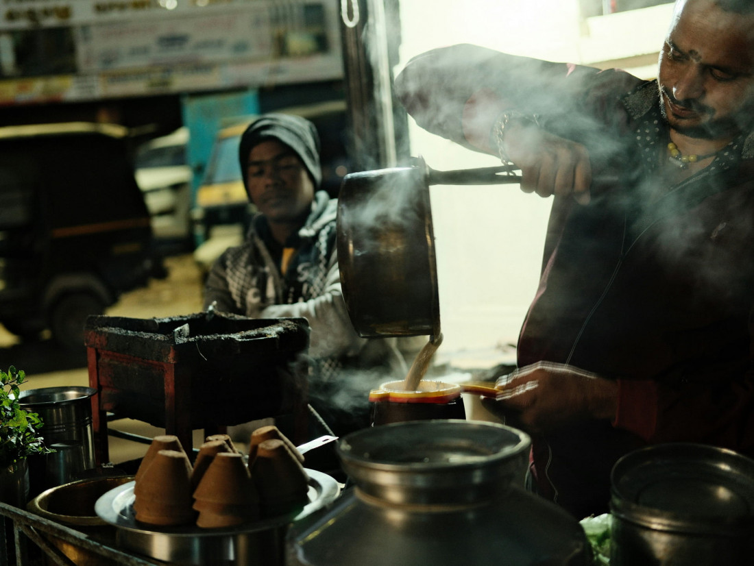 Indian chaiwalla pouring hot masala chai from a height at a night street stall, creating steam. Traditional clay kulhar cups and fresh mint in the foreground. Authentic Indian street food preparation.
