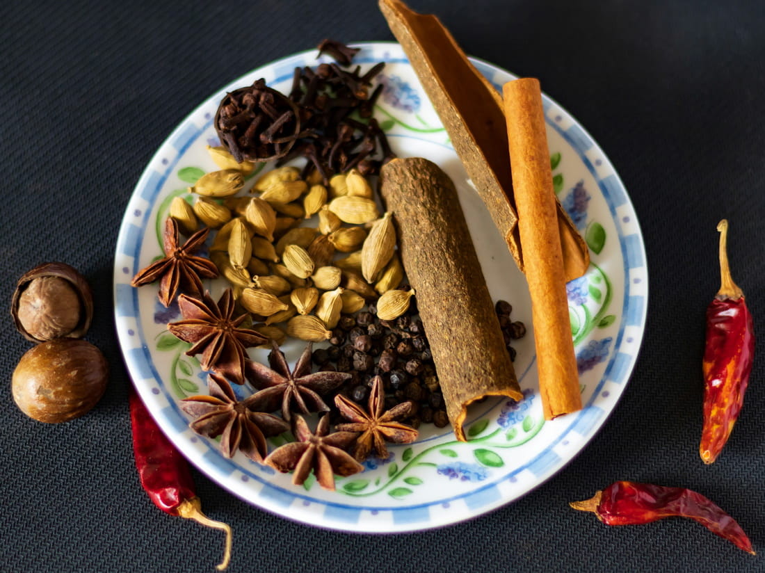 A plate featuring whole spices: cinnamon stick, green cardamom pods, cloves, star anise, and nutmeg.