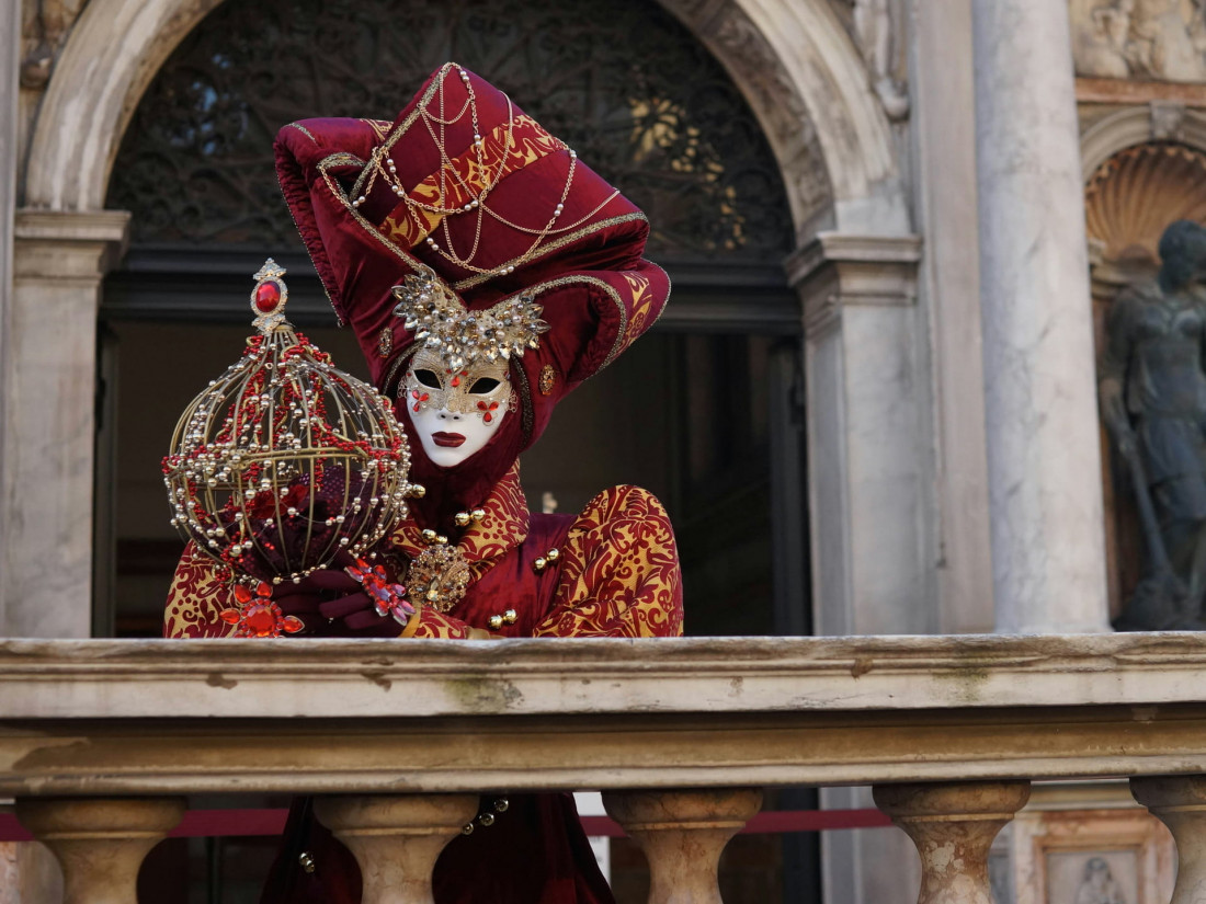 Venetian Carnival participant in an elaborate burgundy velvet costume and gold-embroidered headdress posing with a jeweled cage on a balcony
