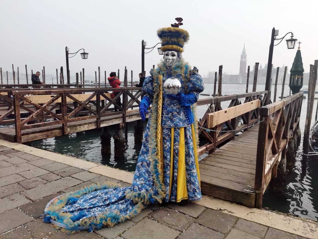 Carnival participant in blue costume holding a disco ball against the lagoon backdrop