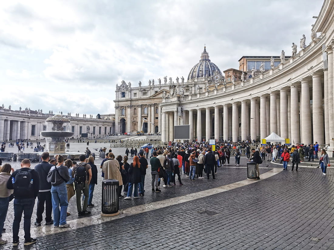 Long queue of tourists waiting to enter St. Peter's Basilica in Vatican City, winding through the square next to the massive colonnade.