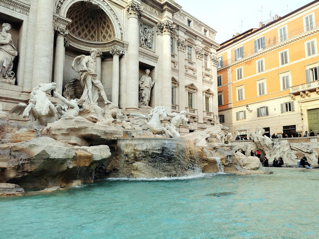 Side angle view of the Trevi Fountain in Rome showing the Oceanus statue and sea horses, with clear turquoise water in the foreground.