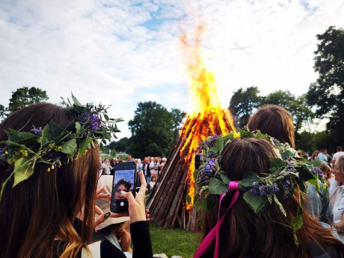 Two women wearing traditional flower crowns taking a smartphone selfie in front of a massive roaring Midsummer bonfire in Estonia.