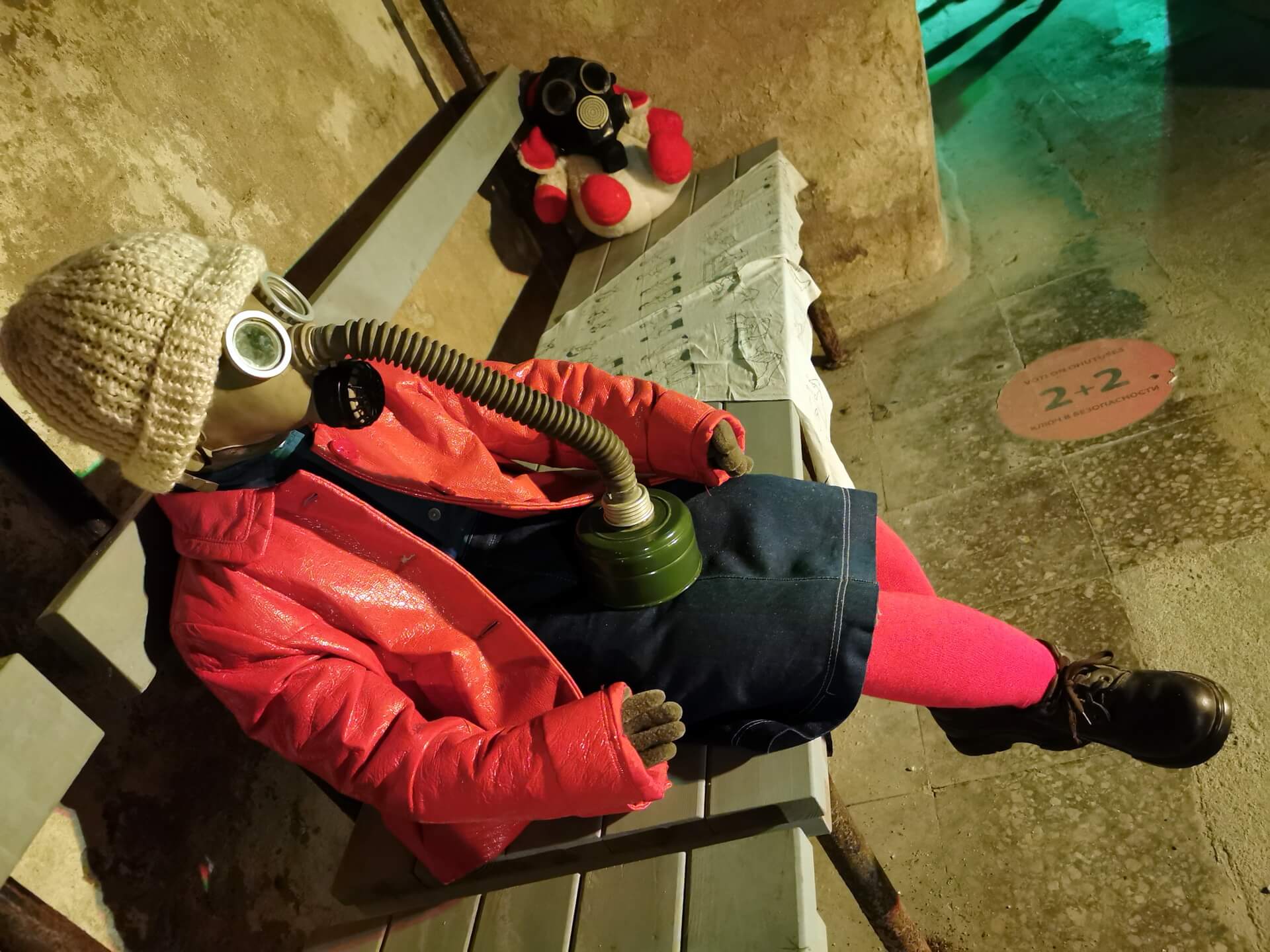 A mannequin wearing a red coat and a Soviet gas mask sitting on a wooden bench in the dimly lit Bastion Tunnels