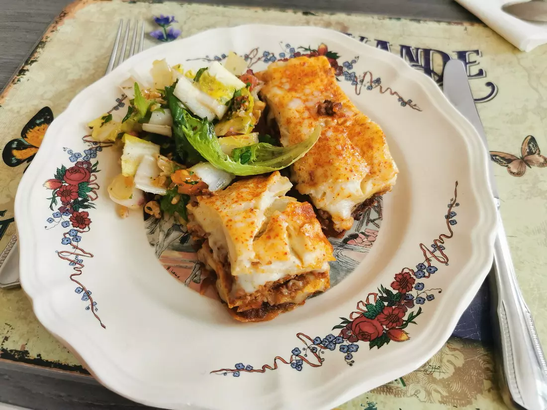 A plate of lasagna with a fresh green salad served on a floral-patterned plate, set on a decorative placemat in a Lyon café.