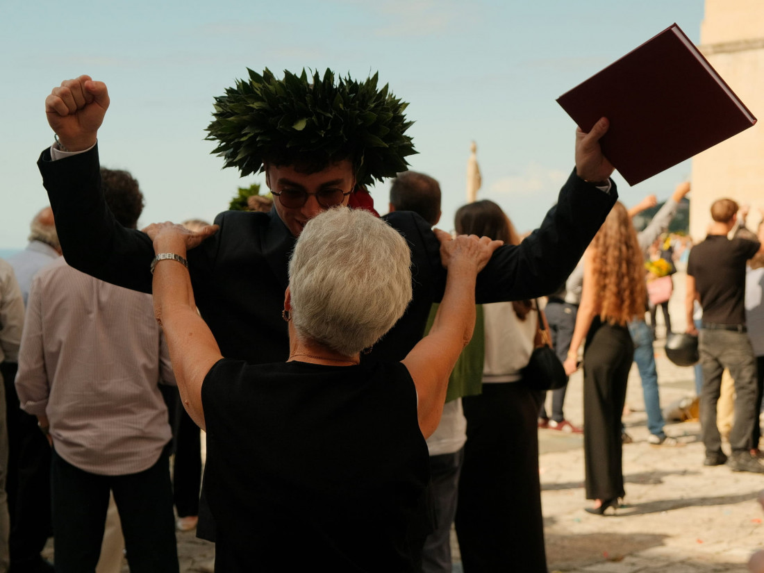 A graduate in a laurel wreath raising his thesis book in celebration, surrounded by family in a public square.