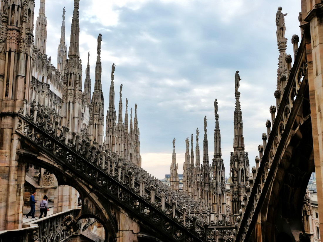 A stunning view of the Milan Duomo rooftop, showcasing intricate Gothic spires and statues under a cloudy sky.