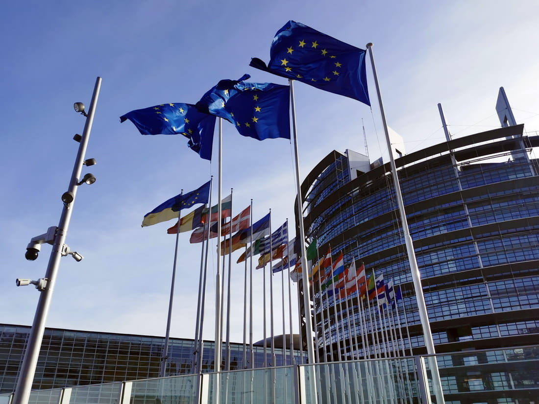 European Union flags flying in front of the Parliament building in Strasbourg against a blue sky.