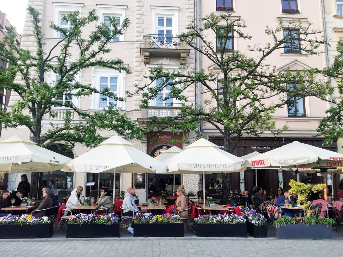 Locals and tourists sitting at outdoor cafe tables under umbrellas on a lively street in Krakow.